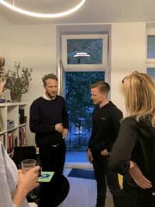 Vier Personen stehen in einem hellen Raum mit Bücherregal und Pflanze, zwei Männer unterhalten sich nahe einer offenen Tür mit Blick auf einen Garten.