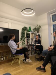 Eine Frau und ein Mann sitzen in einem Wohnzimmer mit Holzfußboden und sprechen miteinander, eine dritte Person ist teilweise sichtbar
